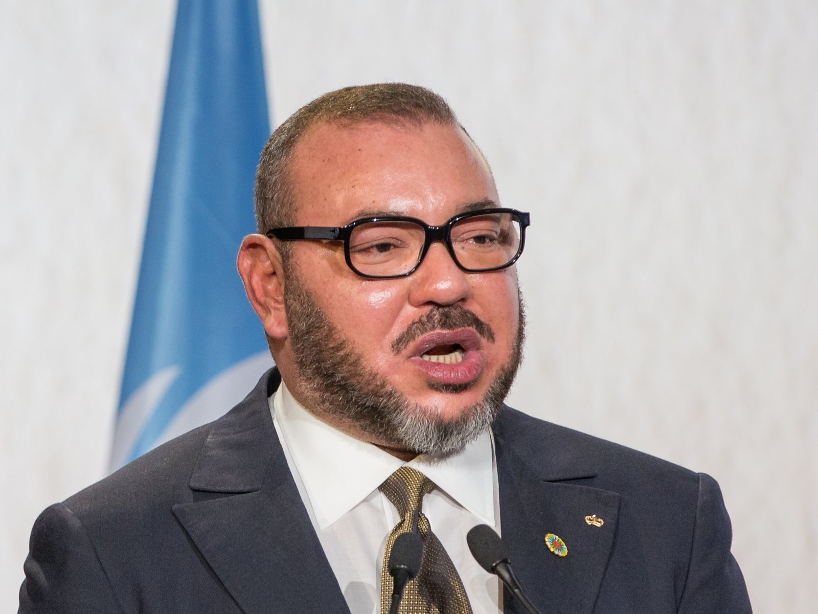 S.M. le Roi Mohammed VI, en costume sombre et lunettes, s'exprimant au pupitre de la COP22 devant le drapeau des Nations unies