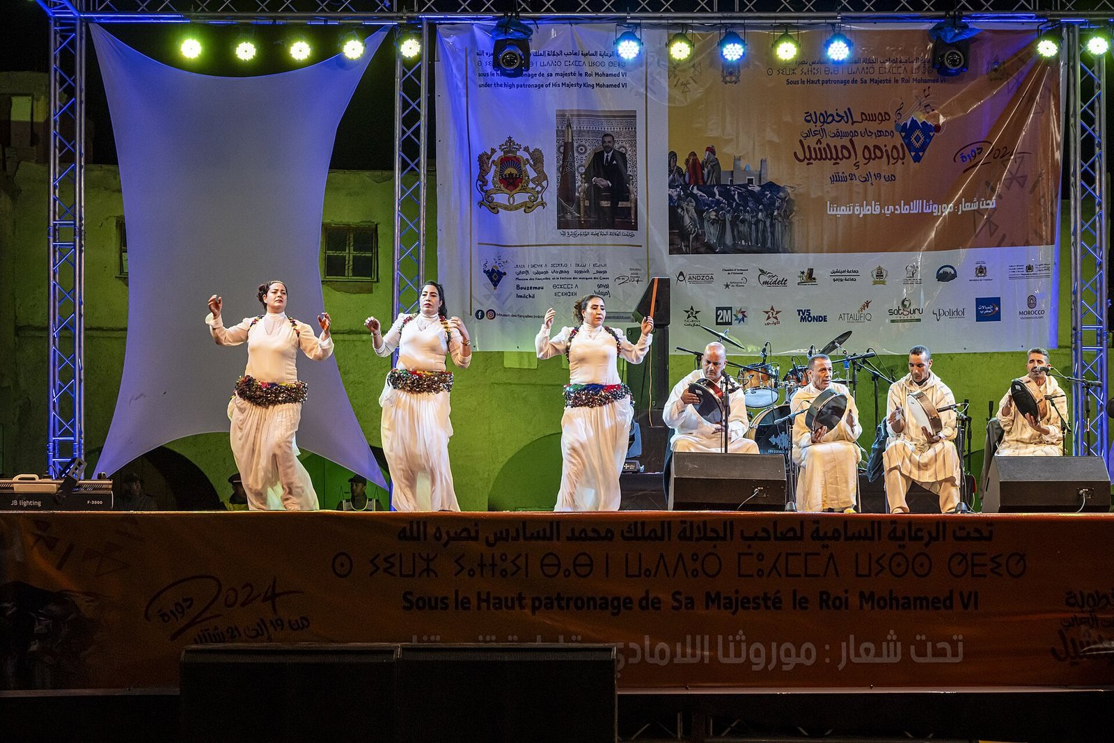 Trois danseuses en robe blanche et quatre musiciens jouant du bendir sur la scène du festival, devant la banderole de l'édition 2024