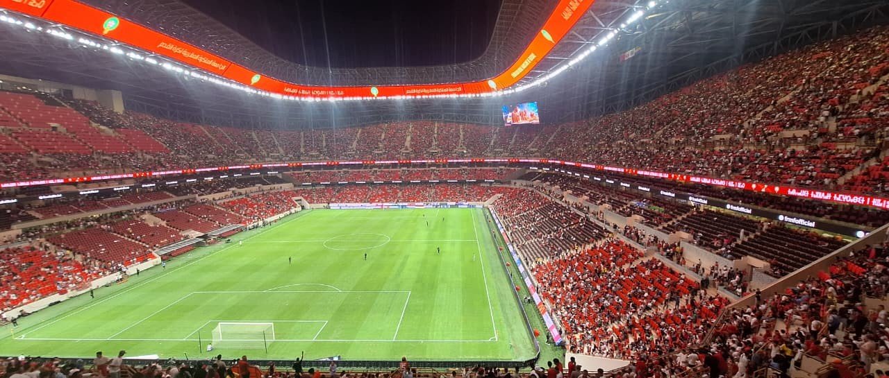 Vue panoramique depuis le haut des tribunes : la pelouse éclairée et des gradins remplis de supporters en rouge