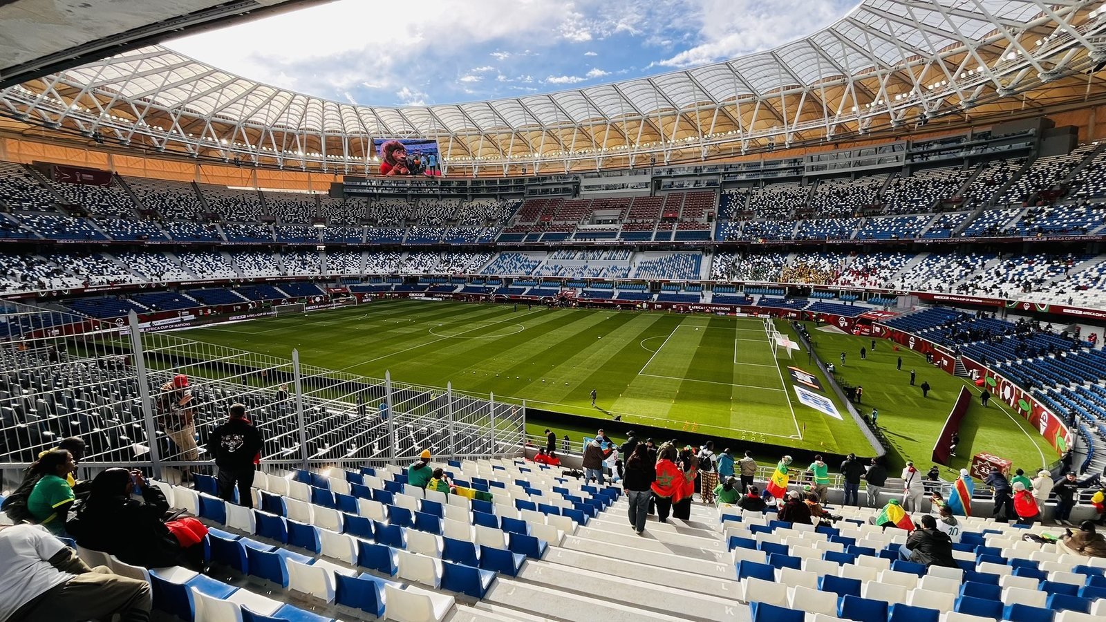 Vue intérieure du Grand Stade de Tanger depuis les gradins : la pelouse, des tribunes aux sièges bleus et blancs encore peu remplies et la toiture qui couvre le stade