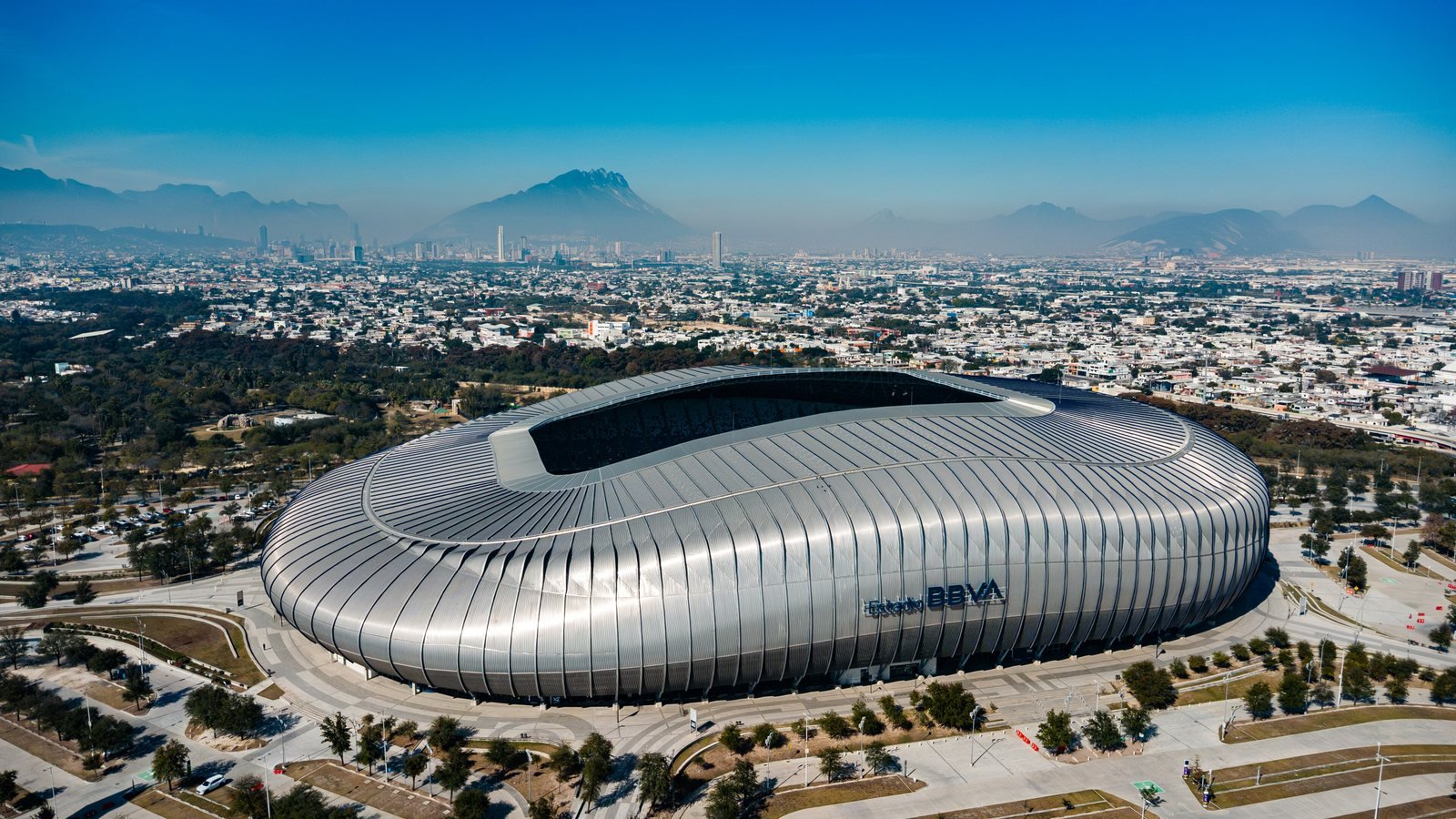 Vue aérienne de l'Estadio BBVA, stade à la façade métallique argentée, avec les montagnes de Monterrey au loin
