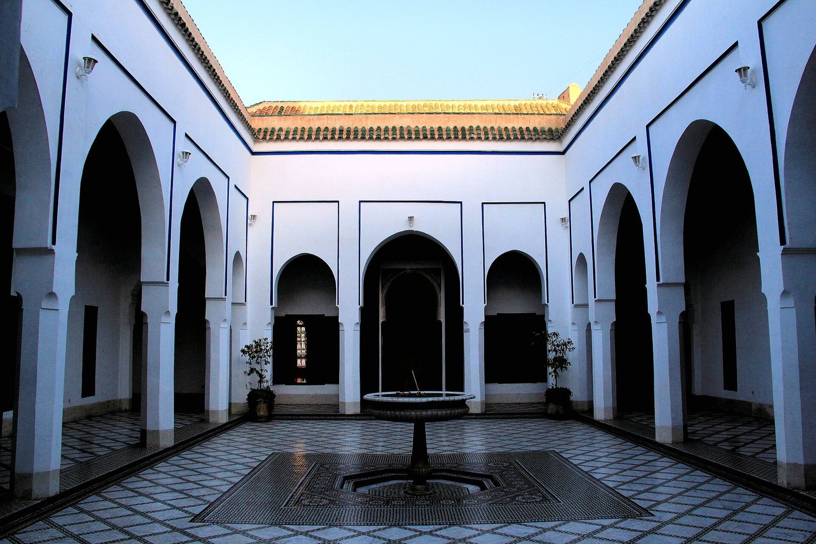Cour du palais Bahia à Marrakech : arcades blanches soulignées de bleu et fontaine au centre d'un sol en zellige
