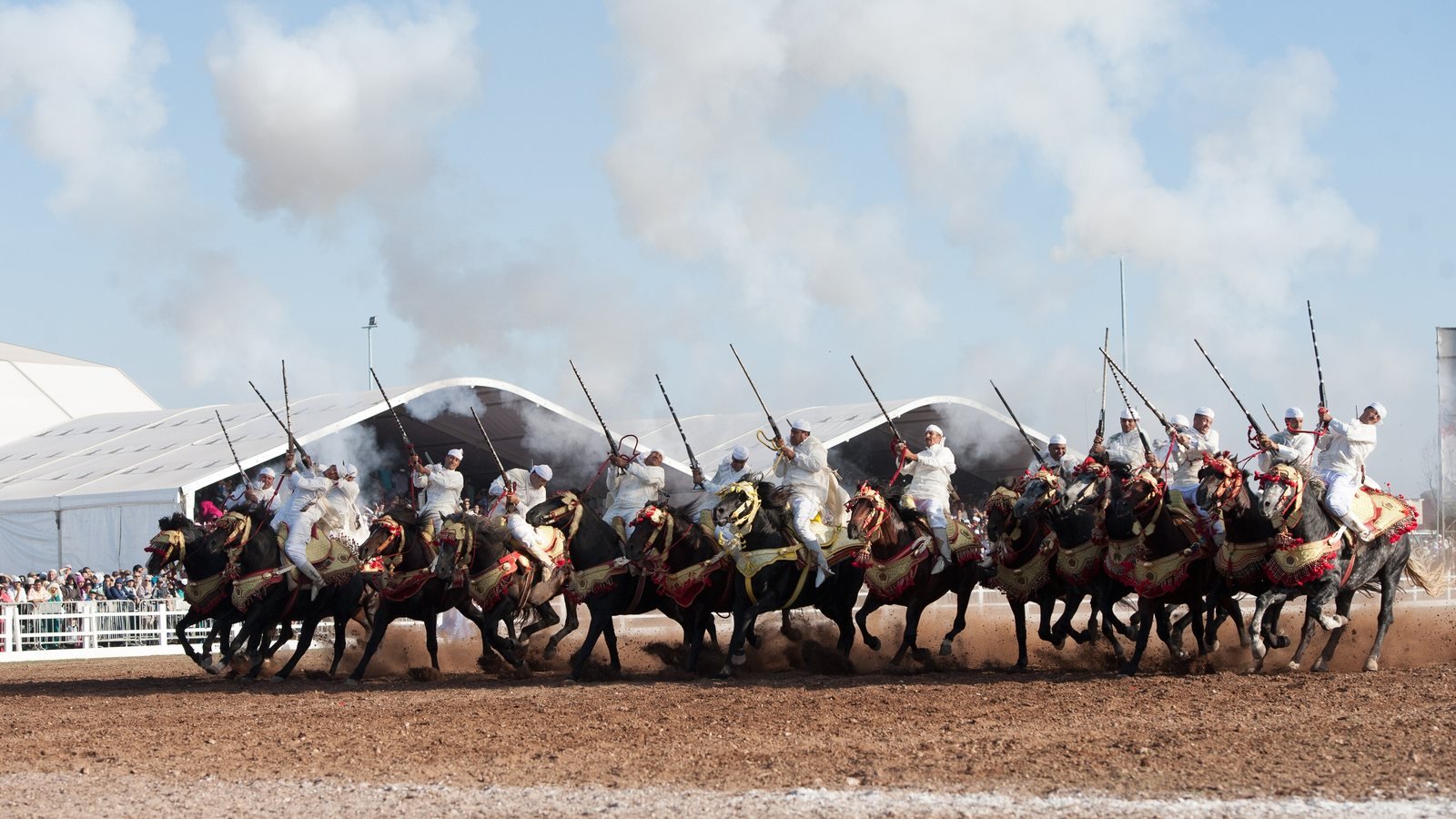 Une sorba de cavaliers en blanc lancée au galop, fusils levés dans la fumée de la poudre, devant les chapiteaux du Salon