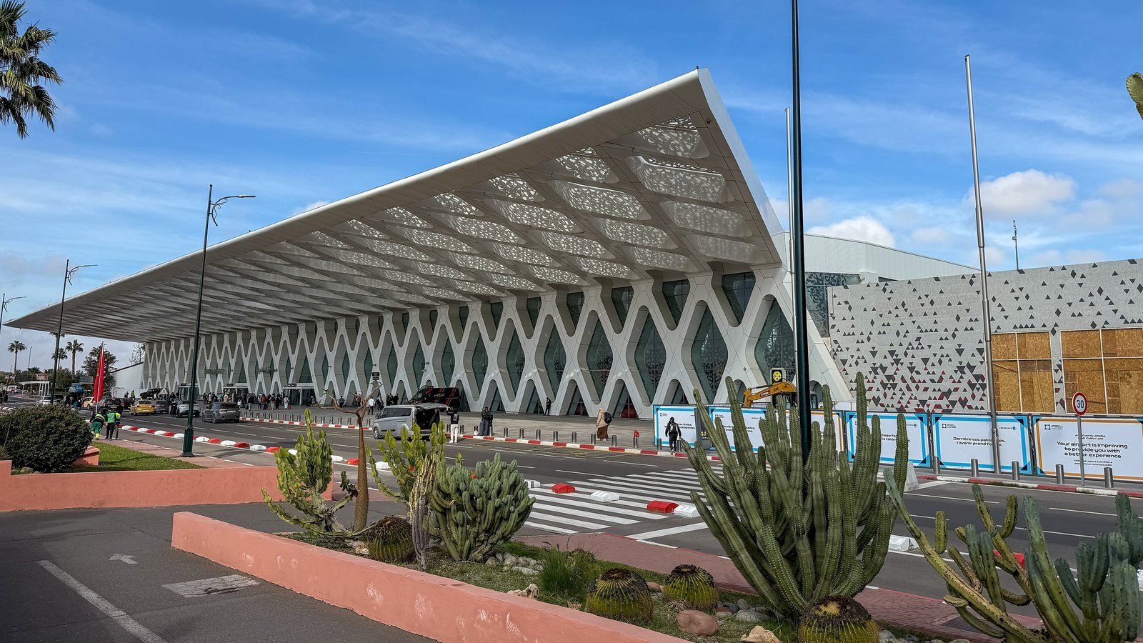 Façade du terminal 1 de l'aéroport Marrakech-Ménara : un auvent blanc ajouré aux motifs géométriques sous un ciel bleu, avec des cactus au premier plan